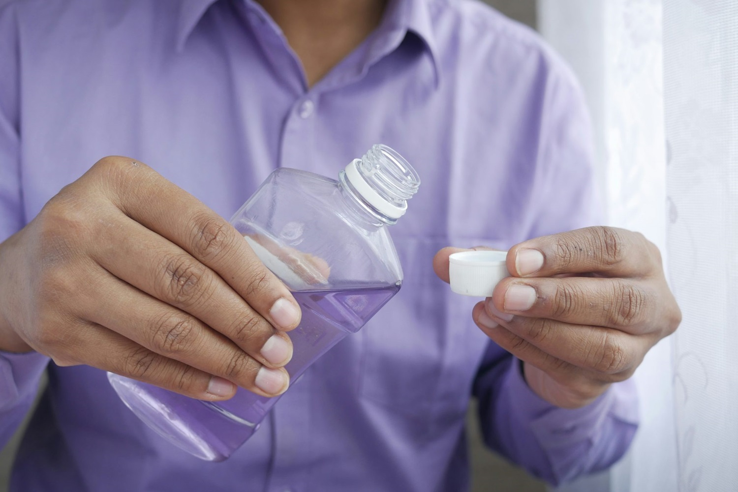 A person in a purple shirt pours purple mouthwash into a bottle cap near a window. - Best Mouthwash